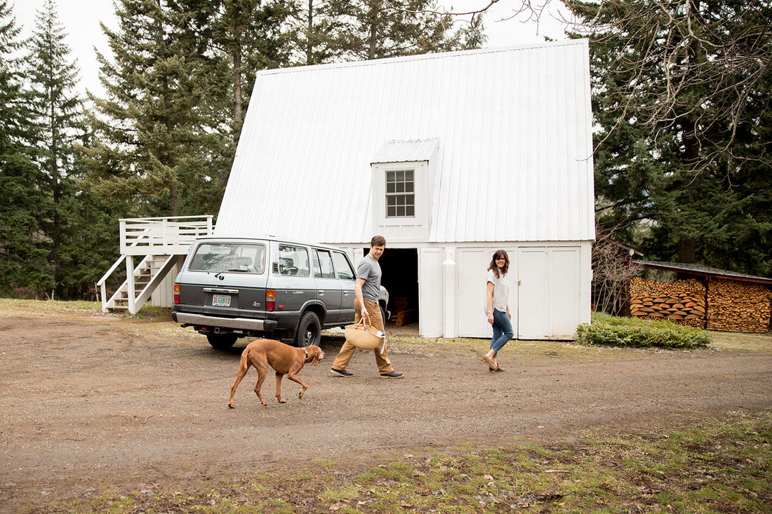 Man carrying a woven basket and woman walking with a brown dog in front of a white A-frame barn and parked SUV in a wooded yard
