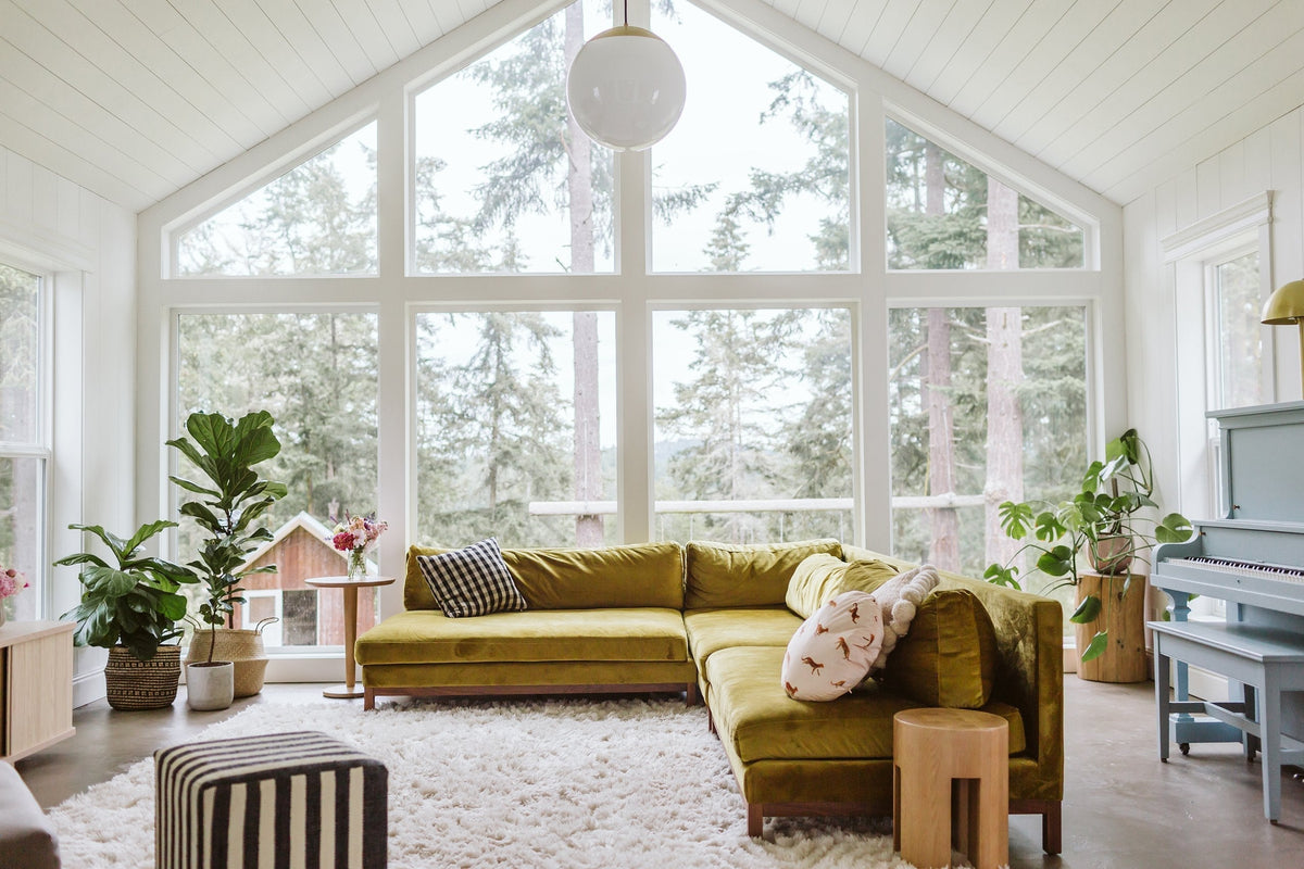 Bright living room with green velvet L-shaped sectional facing floor-to-ceiling windows overlooking evergreen forest, shag rug, potted plants, and a pale blue upright piano