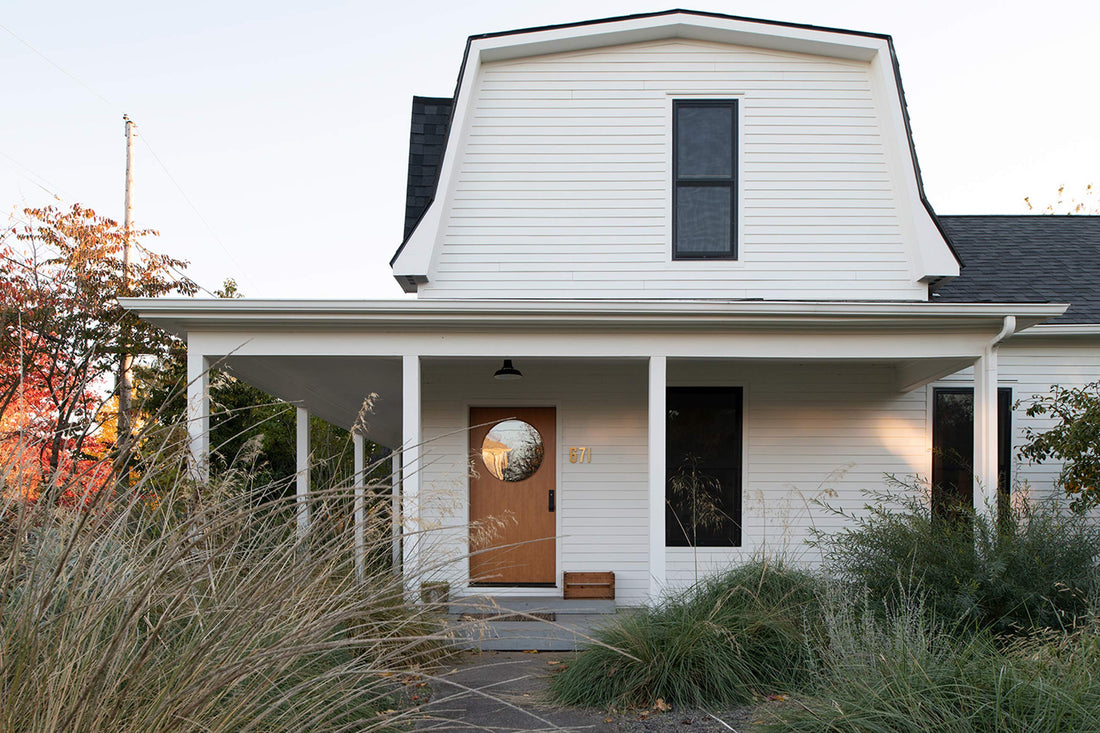 White farmhouse porch with covered entry and black pendant light above a door with round window framed by tall grasses