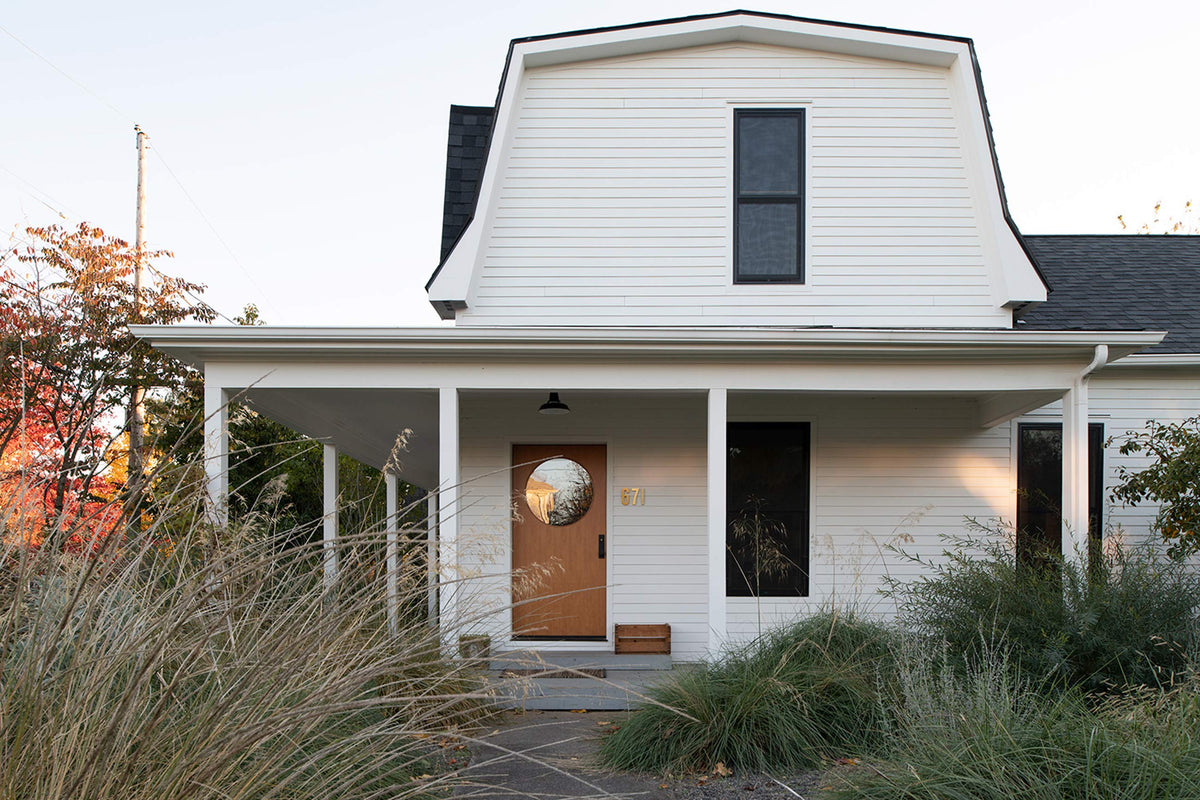 White farmhouse porch with covered entry and black pendant light above a door with round window framed by tall grasses