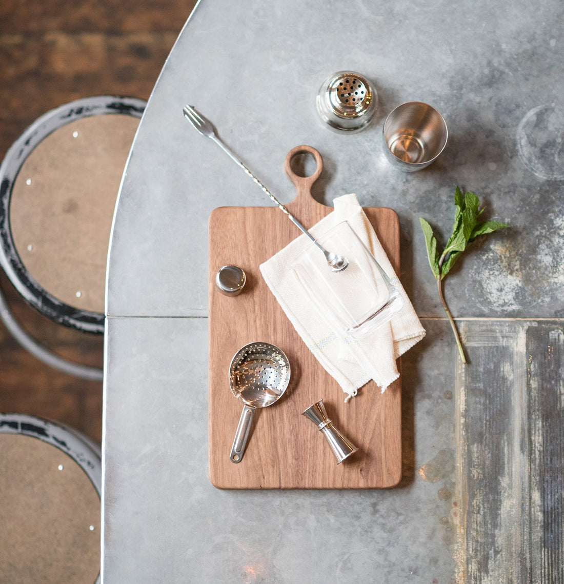 Cocktail tools arranged on a wooden board on a metal table, including a twisted mixing spoon, jigger, strainer, empty glass and a mint sprig