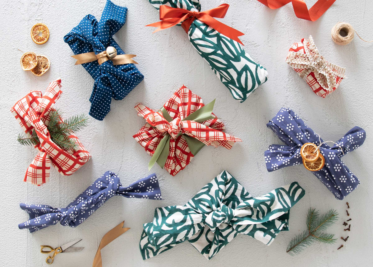 Tenugui cloth wrapped gifts arranged on a light textured surface with ribbons, dried orange slices, scissors, and evergreen sprigs