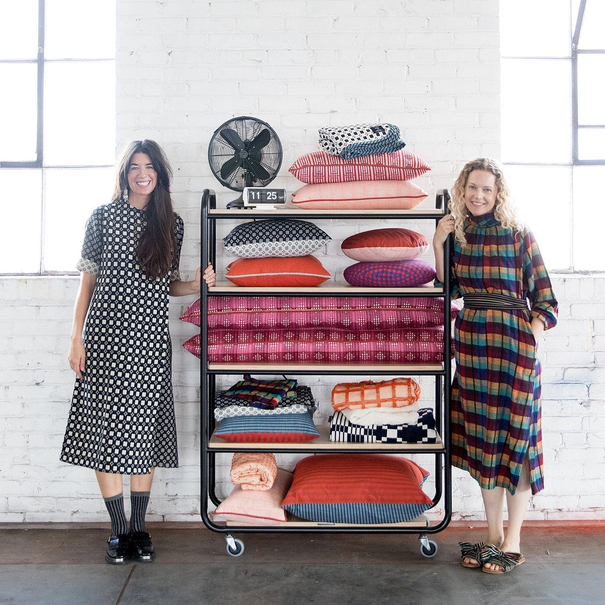 Two women standing beside a rolling shelf stacked with colorful patterned pillows and blankets against a white brick wall