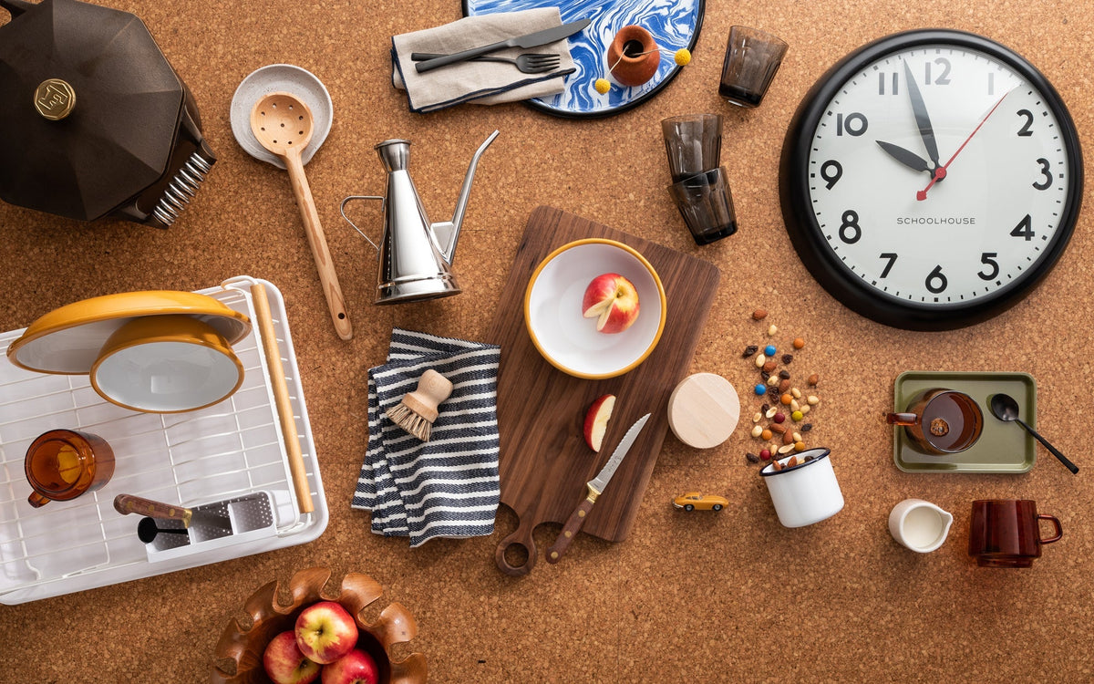 Flat lay of kitchen items on cork: Schoolhouse wall clock, wooden cutting board with sliced apple, yellow-rim bowls, mugs, and utensils