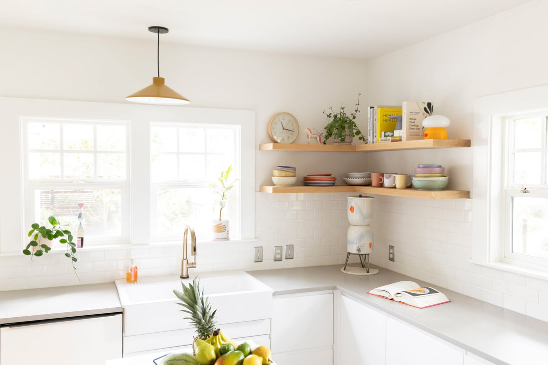Sunlit white kitchen with farmhouse sink and brass faucet, wooden floating shelves styled with colorful dishes, plants, and cookbooks