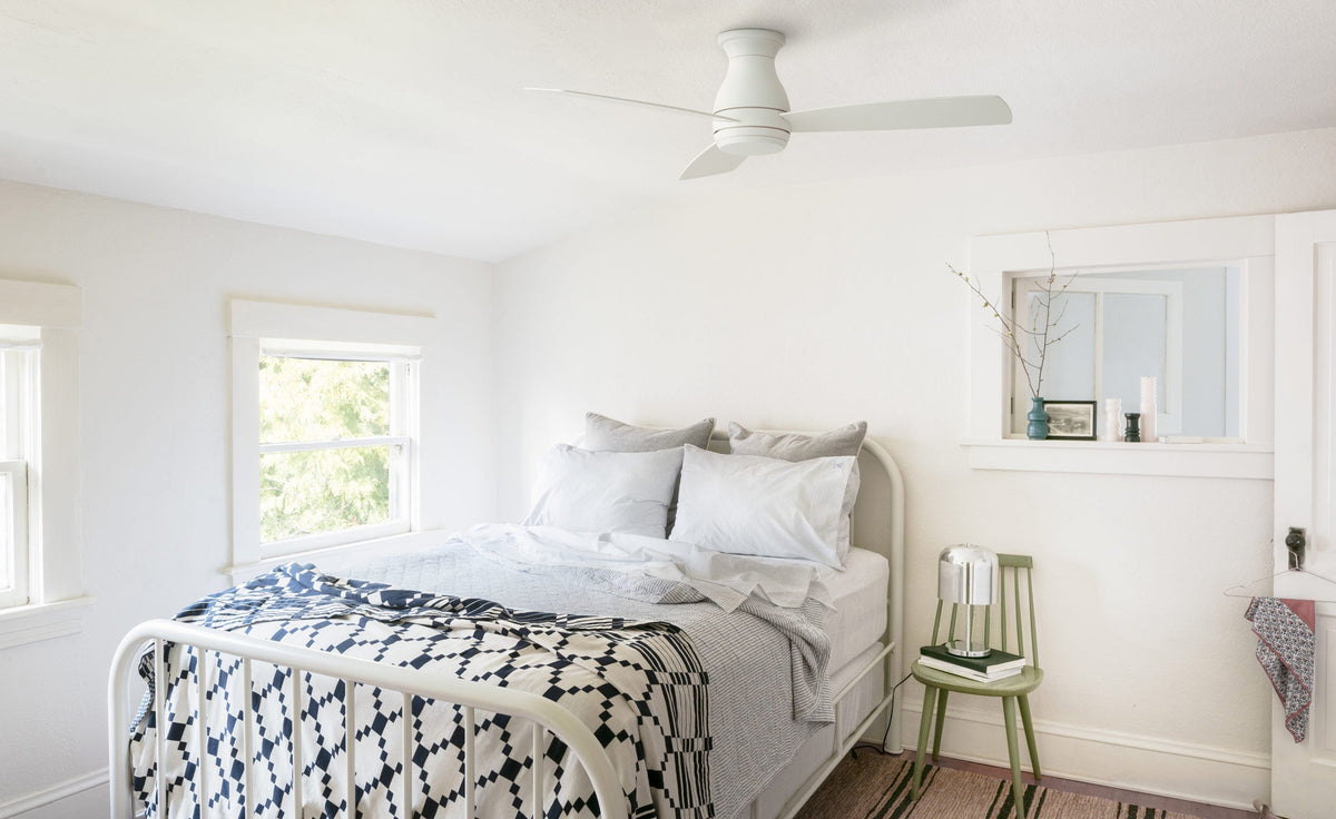 White ceiling fan above a bright bedroom with a metal bed, gray pillows, navy-and-white patterned blanket and green chair