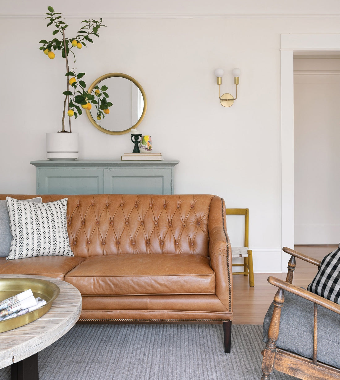 Brown leather tufted sofa with patterned pillow, round brass mirror, potted lemon tree on blue console, brass wall sconce