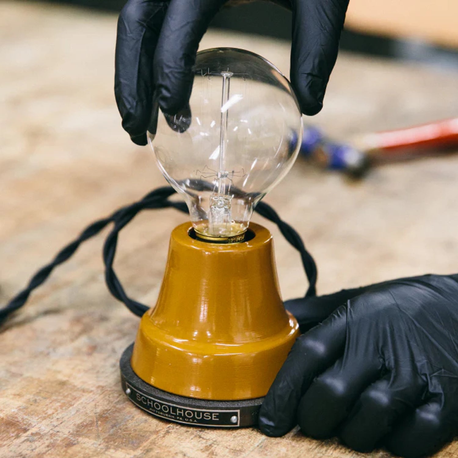 Technician wearing black gloves inserts a clear globe bulb into a mustard-yellow lamp base on a wooden workbench