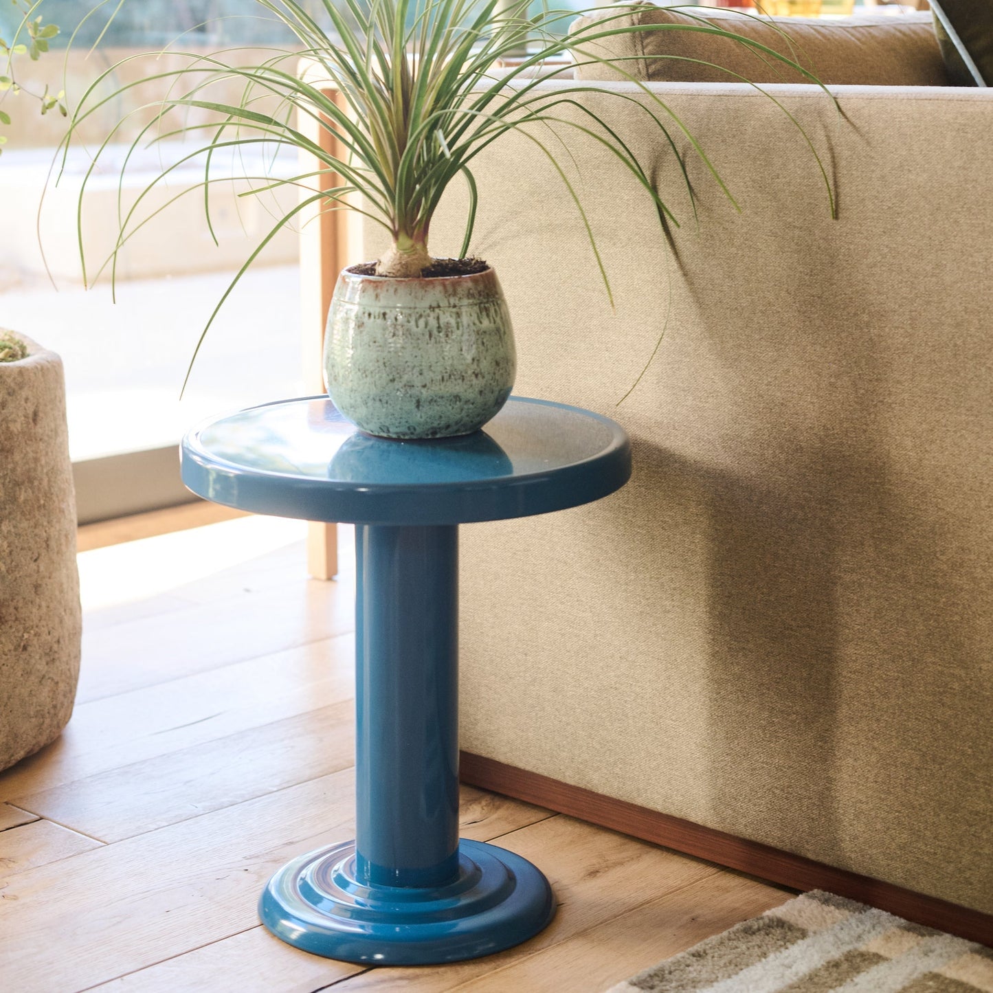 Side view of Owen Side Table showing glossy pool-blue pedestal base and round top with a potted plant beside a beige sofa
