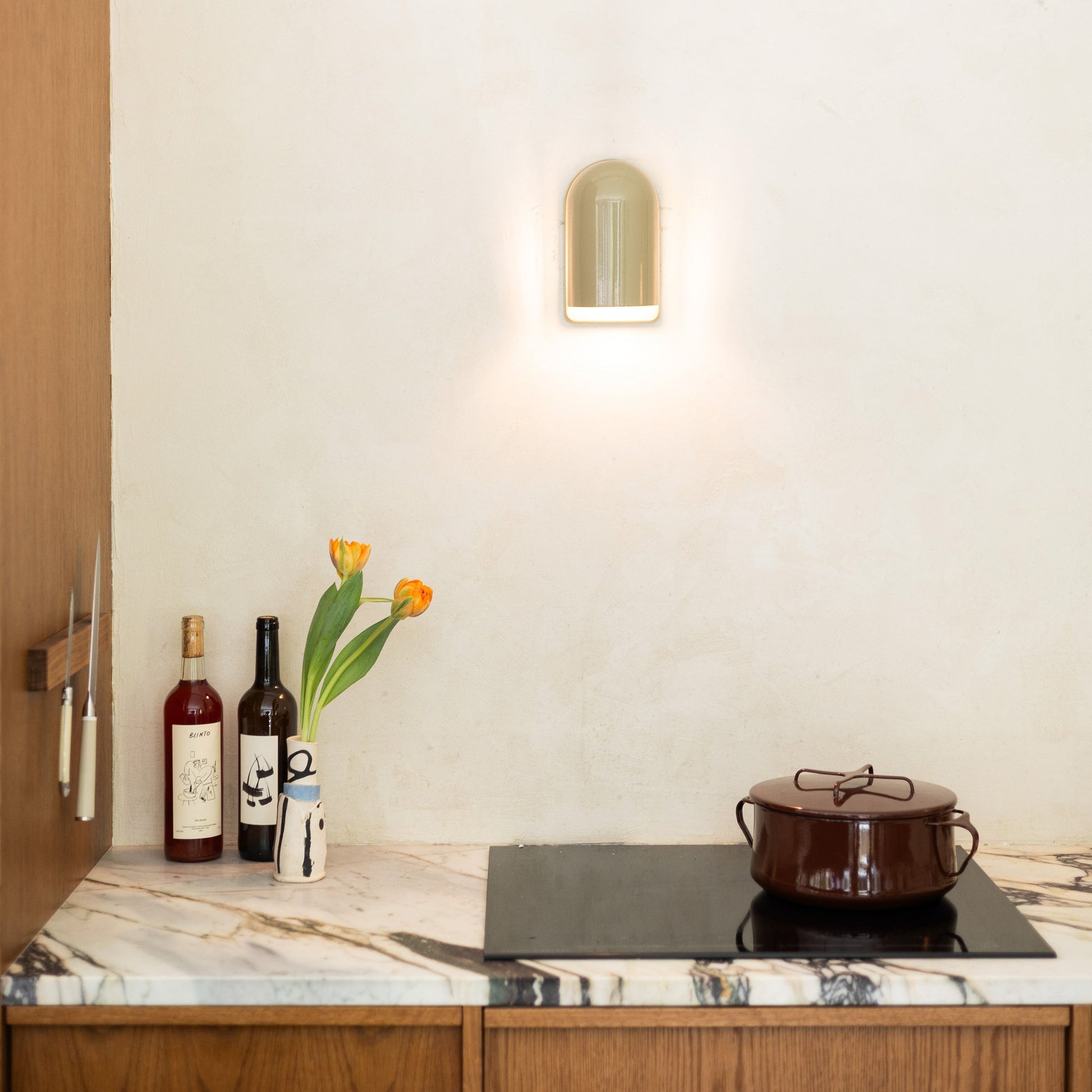 Allegheny Slim Sconce casting warm light on a neutral kitchen wall above a marble countertop with tulips and cookware