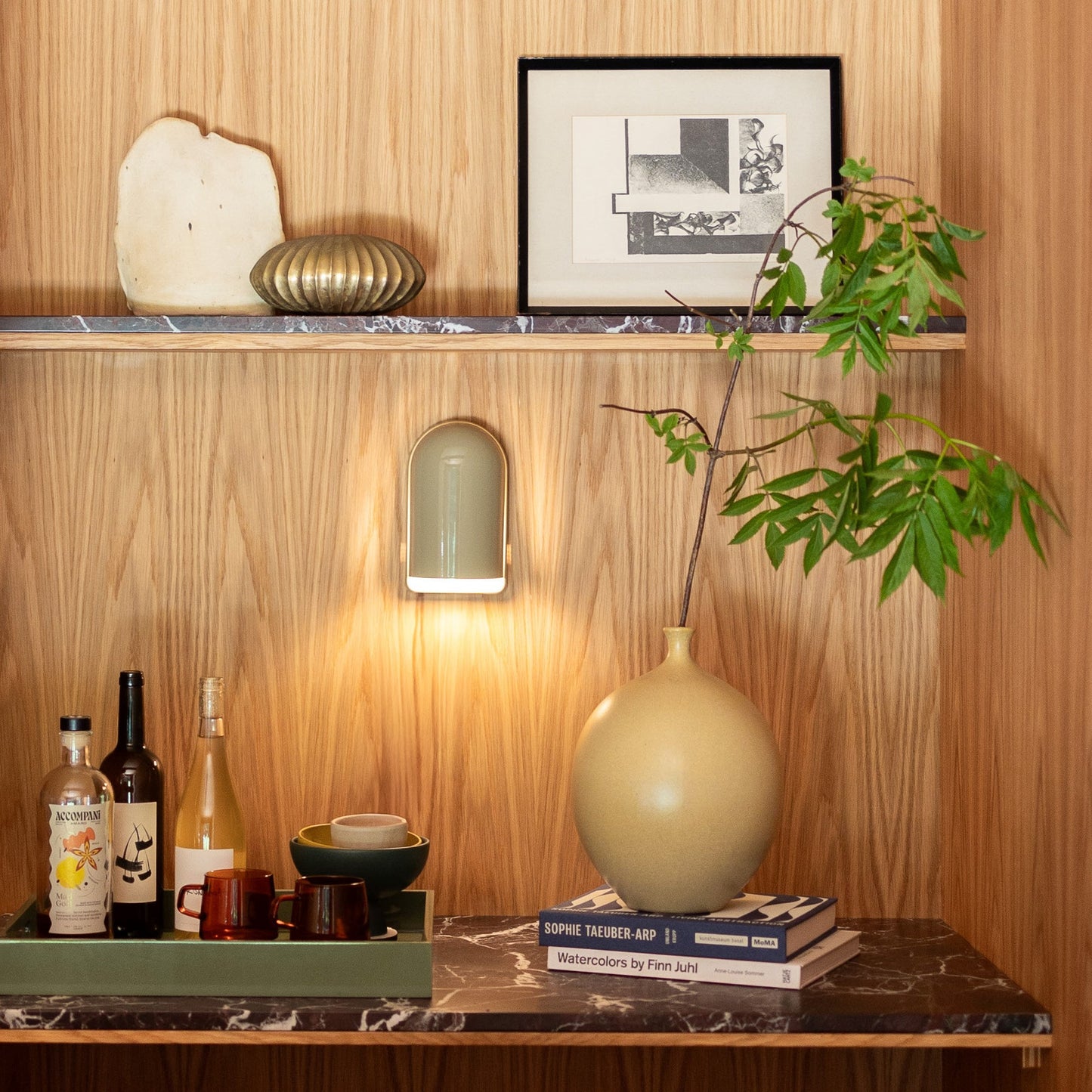 Allegheny Slim Sconce lit on a wood-paneled wall above a marble shelf styled with framed art, pottery, brass accent, and a potted branch