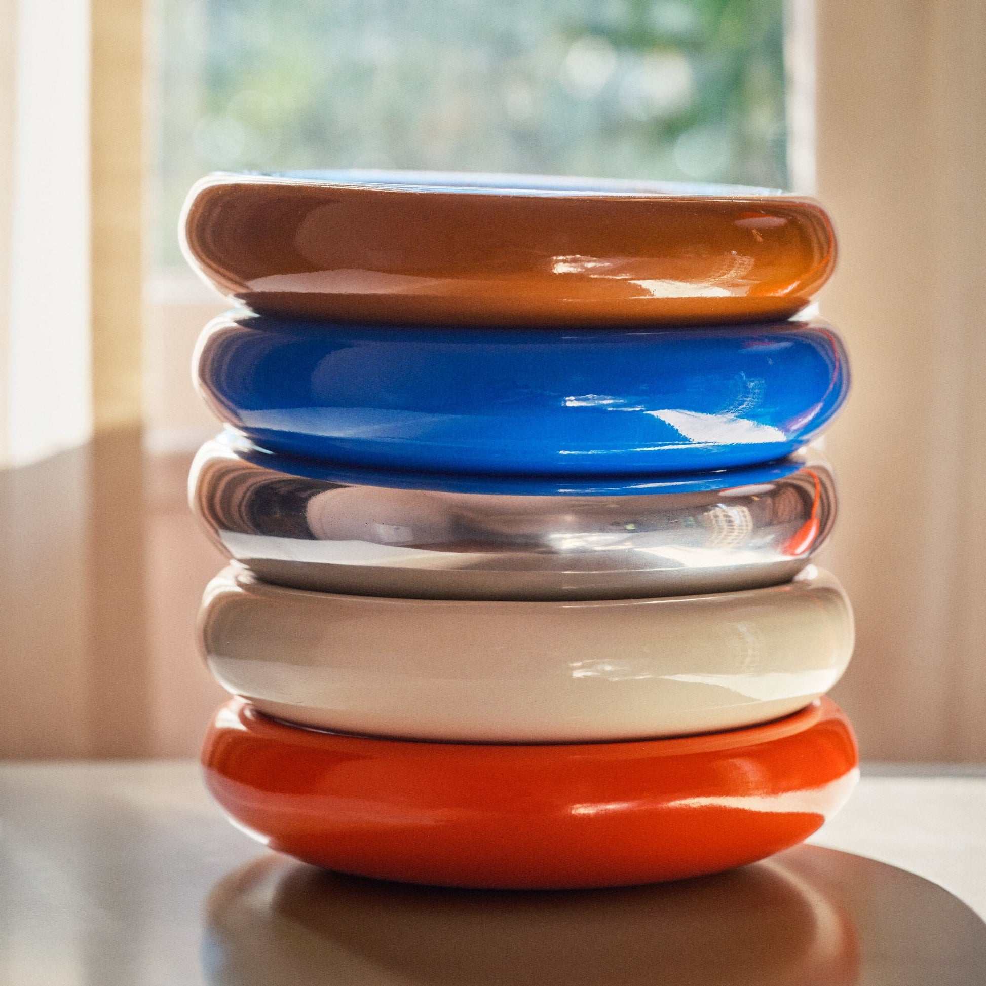 Close-up side view of stacked Yves cast metal catch-all dishes showing glossy finish and color variations against a blurred window background