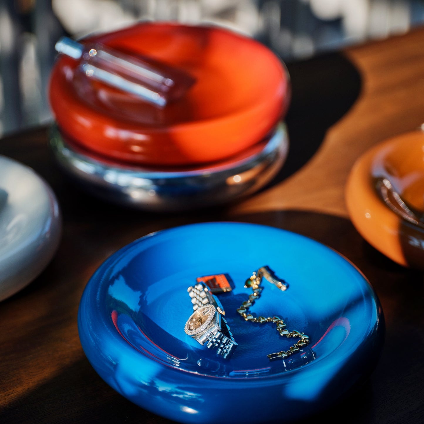 Close-up of blue Yves cast metal catch-all holding a watch and chain, with blurred red and orange bowls on a wooden table