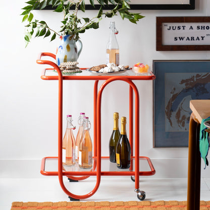 Three-quarter view of Lolly Bar Cart showcasing bent steel frame, tempered glass shelves, and caster wheels against a white wall