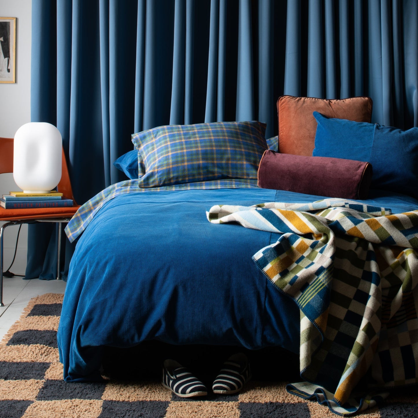 Velvet piped pillows arranged on a bed with blue bedding, plaid throw, rust velvet pillow with contrast piping, and deep blue curtains