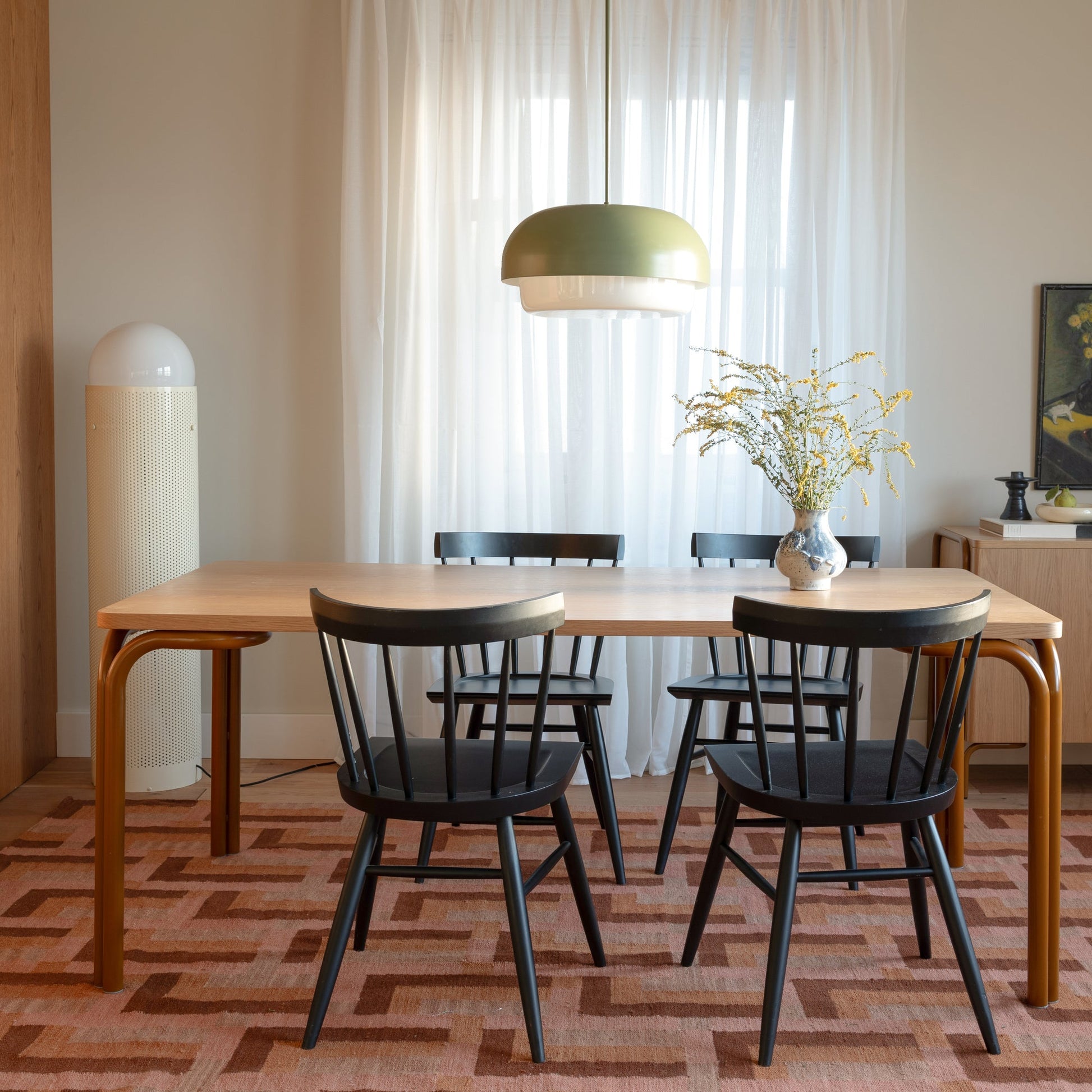 Straight-on view of Forma Dining Table showcasing light oak top and rounded tubular legs with black chairs and sheer curtains in a dining room