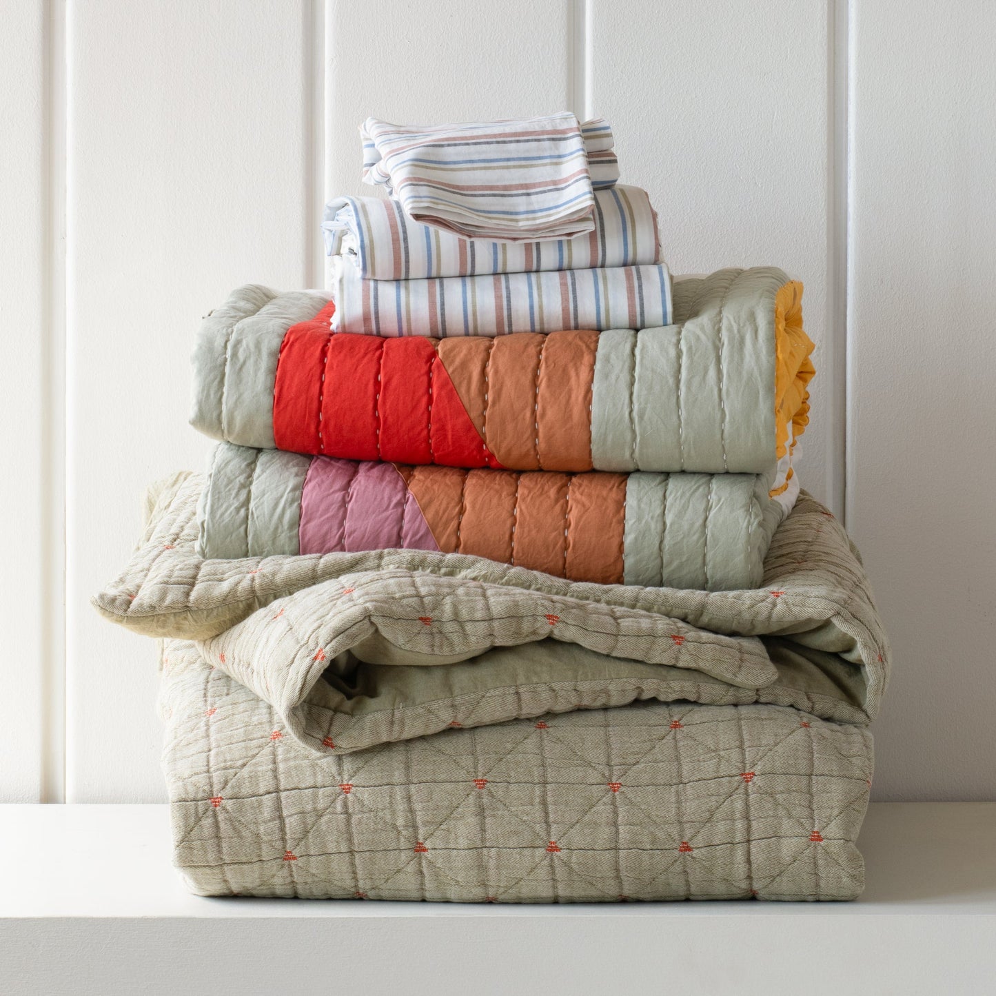Front view of Peak Stitch Duvet showing triangular quilting and red stitch accents with folded striped sheets on a white wall