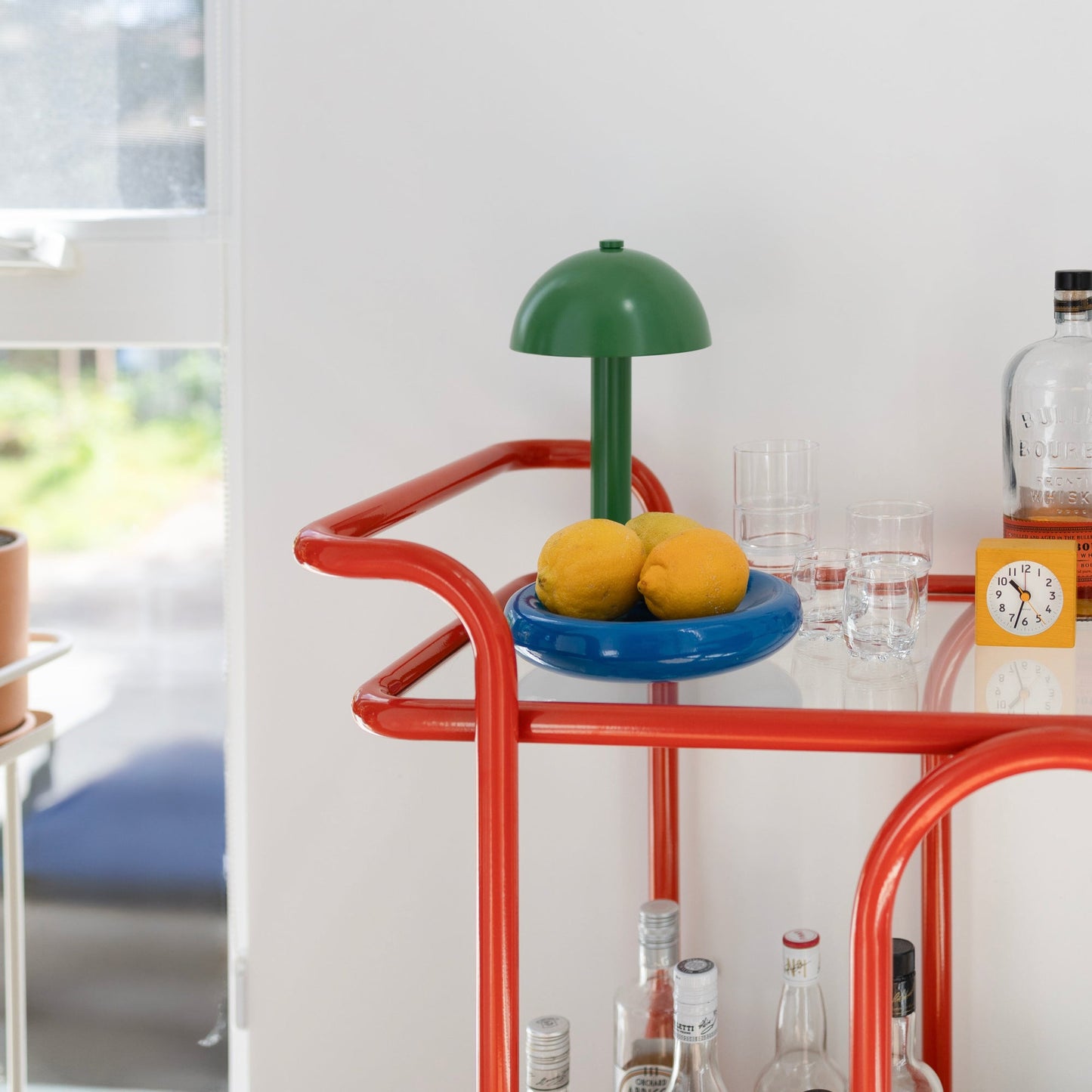 Three-quarter view of Lolly Bar Cart with red bent-steel frame, green lamp, and lemons and glassware on white background