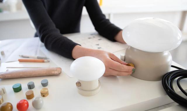 Person adjusting two mushroom-shaped table lamps on a white workbench with color sample knobs and sketches
