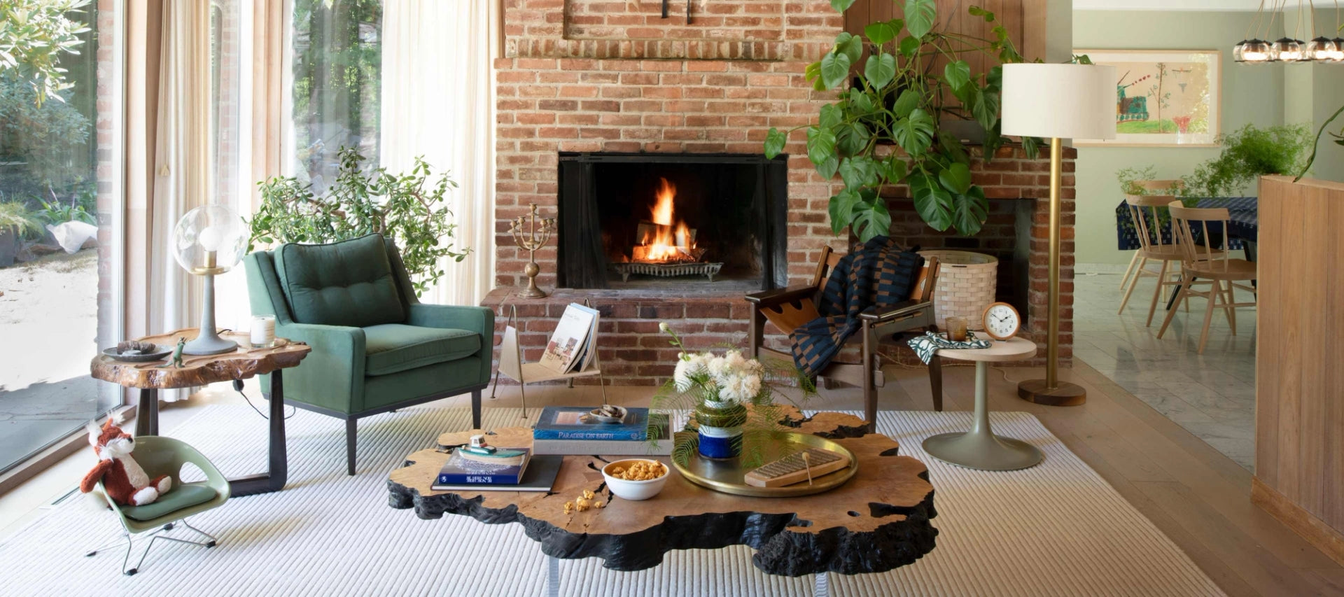 Cozy living room with a lit brick fireplace, green armchair, wood-slab coffee table, plants, and a view into a dining area