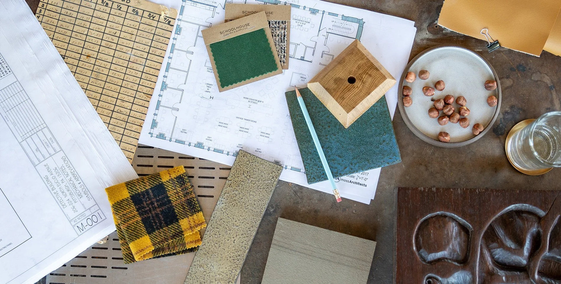 Flat lay of fabric and material samples, a wood block, pencil, plate of hazelnuts, and architectural floorplans on a work table