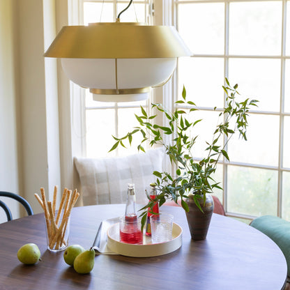 Front close-up of Alfred Pendant showing natural brass shade and white opal glass diffuser hanging over a dining table by sunlit windows