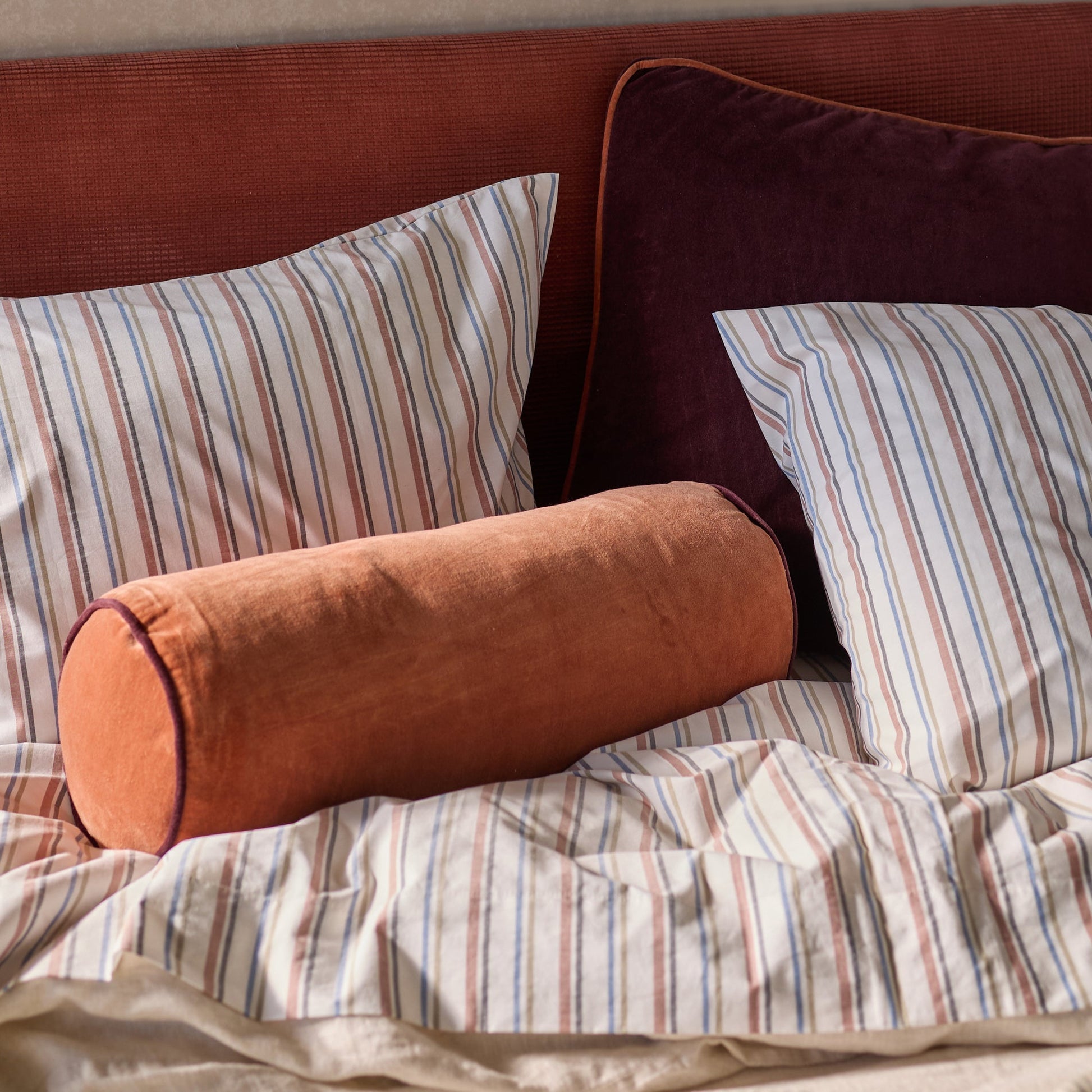 Close-up of Velvet Piped Pillow showing raisin velvet with rust piped edge on bed with striped shams and rust headboard