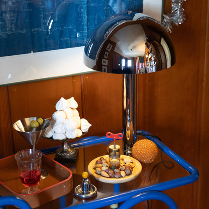 Three-quarter view of Lolly Bar Cart, cobalt gloss, showing bent metal frame and tempered glass shelf styled with a chrome lamp and snacks
