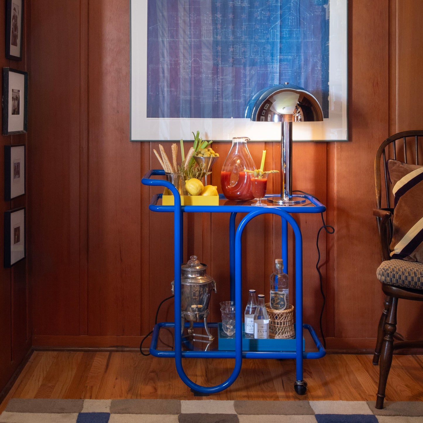 Front view of Lolly Bar Cart showcasing cobalt glossy bent-steel frame, two glass shelves, and caster wheels against a wood-paneled wall