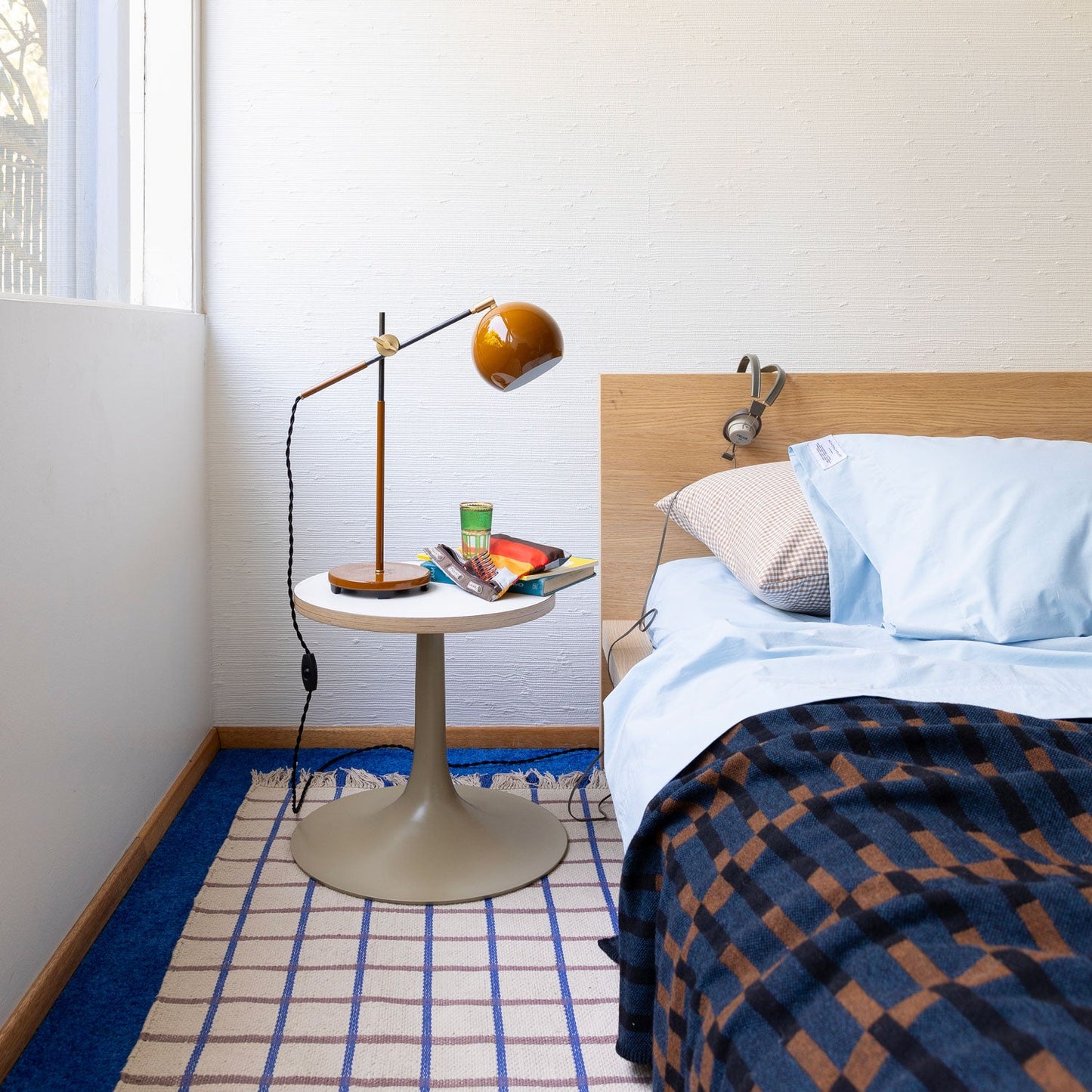 Bedside scene with light blue pillowcase and small gingham pillow on a wooden headboard, round side table with amber desk lamp, books, and a plaid throw