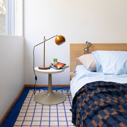 Bedside scene with light blue pillowcase and small gingham pillow on a wooden headboard, round side table with amber desk lamp, books, and a plaid throw