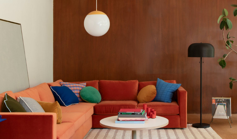 Living room with orange-red sectional sofa and colorful pillows, round coffee table with books, globe pendant and floor lamp