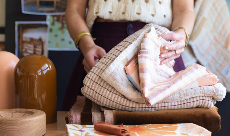 Person folding stacked patterned fabrics on a wooden table with amber ceramic vases and a wooden bowl in a studio