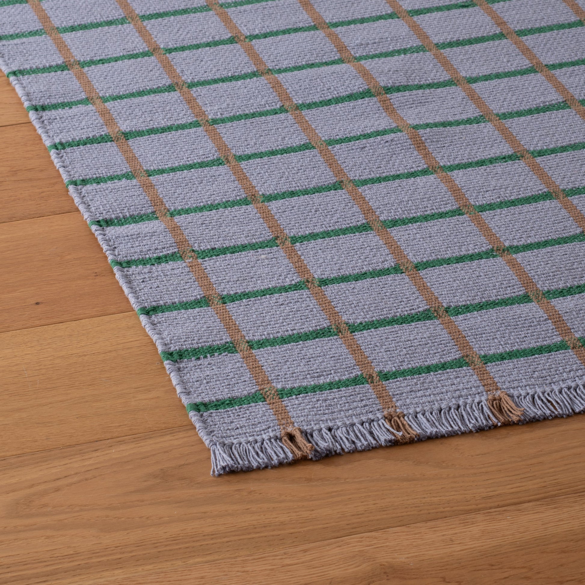 Angled close-up of Cotton Grid Flatweave Rug showing light-gray weave with green and tan grid stripes and fringe on wood floor