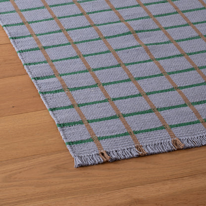 Angled close-up of Cotton Grid Flatweave Rug showing light-gray weave with green and tan grid stripes and fringe on wood floor