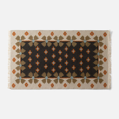 Top-down view of Flora Wool Flatweave Rug with folk floral pattern, dark center and cream border, rust and olive accents on light gray background