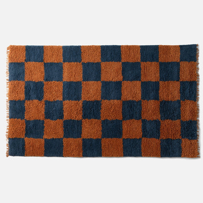 Top edge of Jacques shag wool rug showing rust and navy checked high-pile wool, visible fringe on white background
