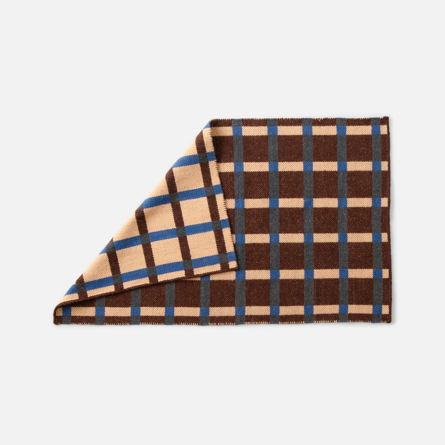 Angled close-up of Plaid Wool Reversible Rug showing folded corner revealing reversible tan, brown, blue and gray plaid on white background