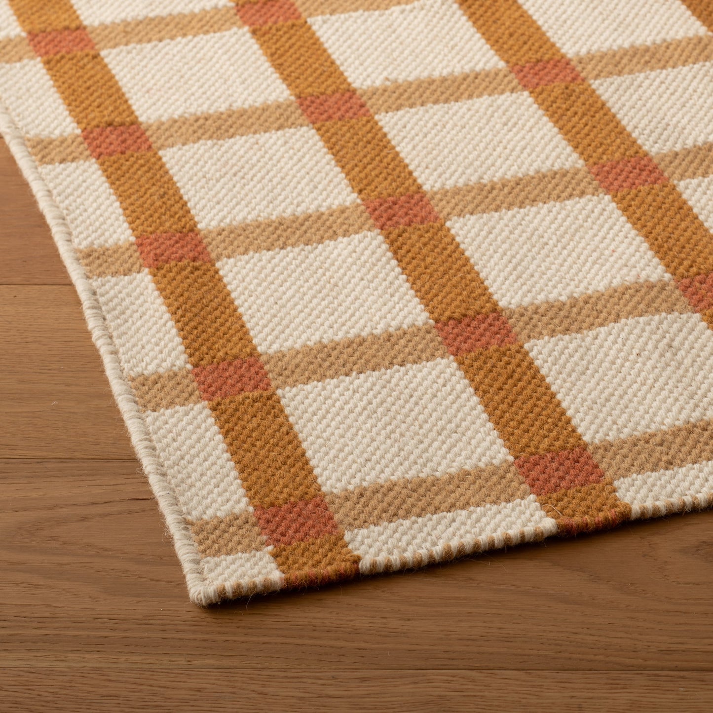 Angled close-up of Plaid Wool Reversible Rug showcasing handwoven natural-dyed wool plaid in cream, tan, mustard, and rust on a wood floor