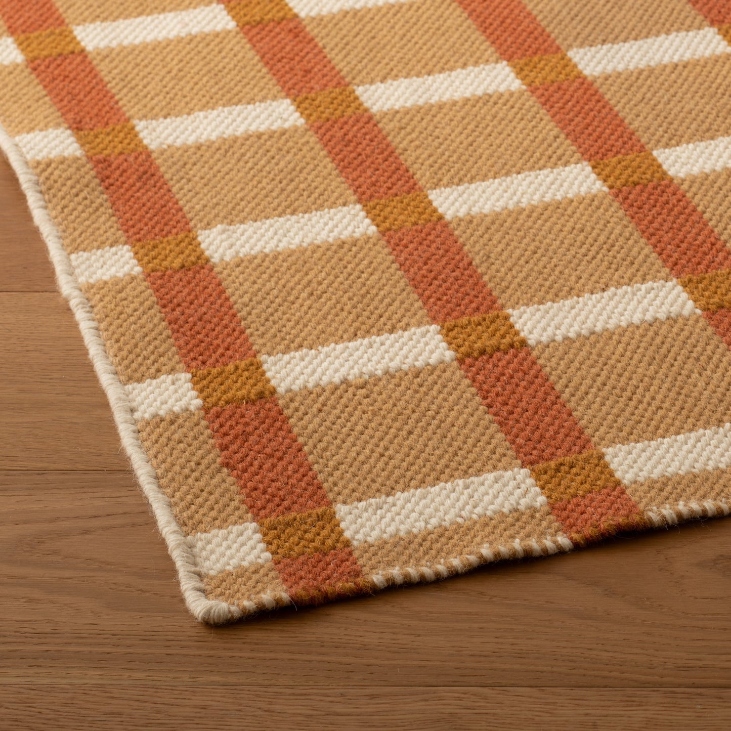 Close-up of Plaid Wool Reversible Rug showing handwoven texture, tan/cream/rust plaid and finished edge on wood floor
