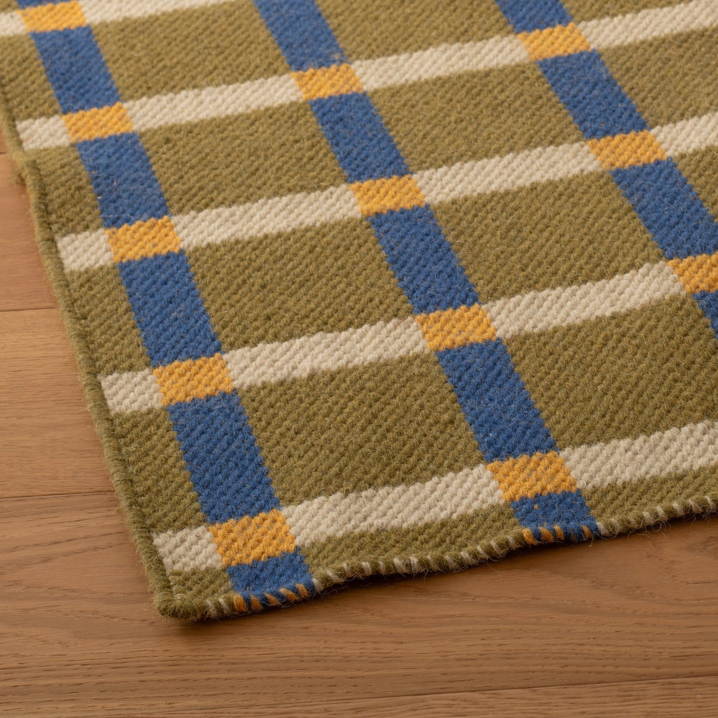 Angled close-up of Plaid Wool Reversible Rug showing handwoven wool texture and olive, cream, blue, and yellow plaid on a wood floor