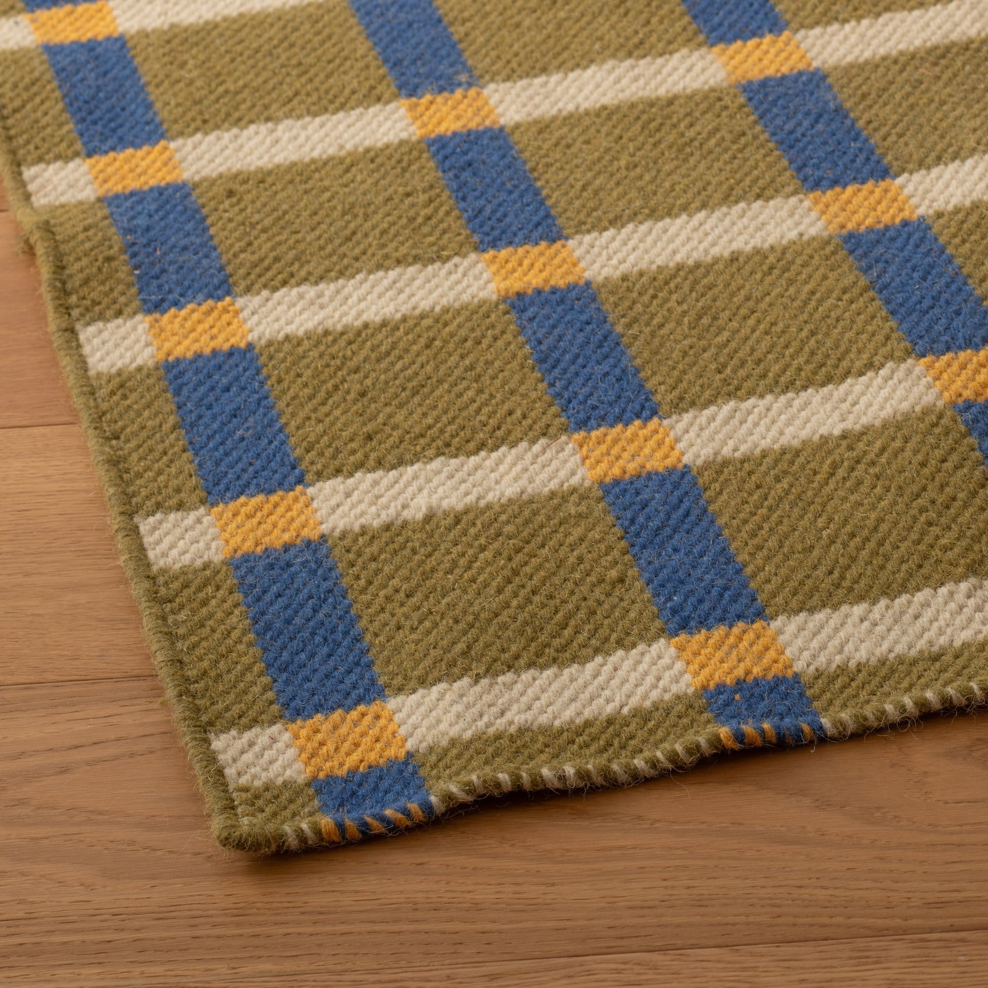Angled close-up of Plaid Wool Reversible Rug showing handwoven wool texture and olive, cream, blue, and yellow plaid on a wood floor
