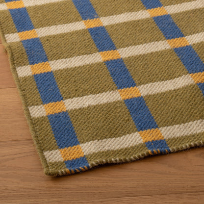 Angled close-up of Plaid Wool Reversible Rug showing handwoven wool texture and olive, cream, blue, and yellow plaid on a wood floor