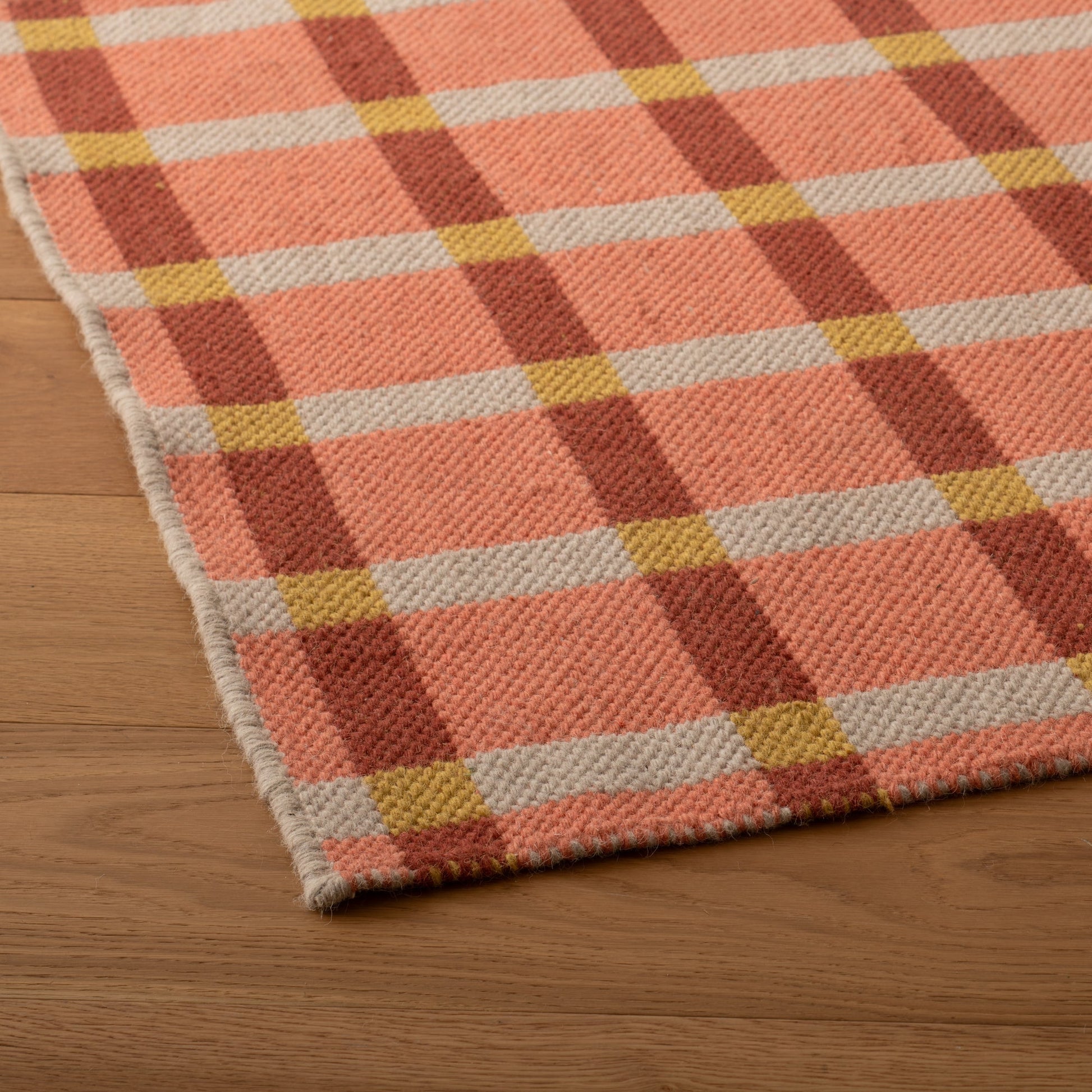 Close-up of Plaid Wool Reversible Rug corner showing handwoven plaid in pink, rust, cream and mustard on wood floor