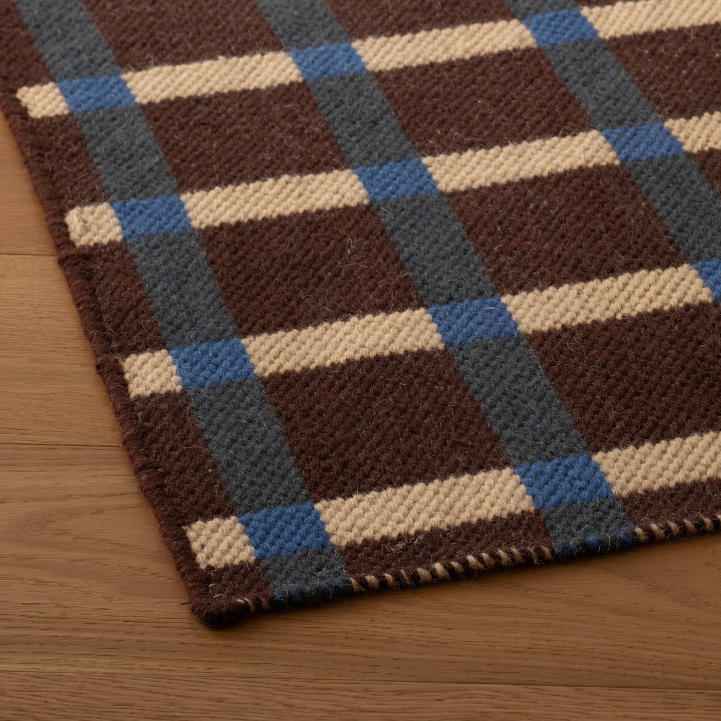 Close up of Plaid Wool Reversible Rug showing handwoven texture, brown cream and blue plaid and whipstitched edge on wood floor