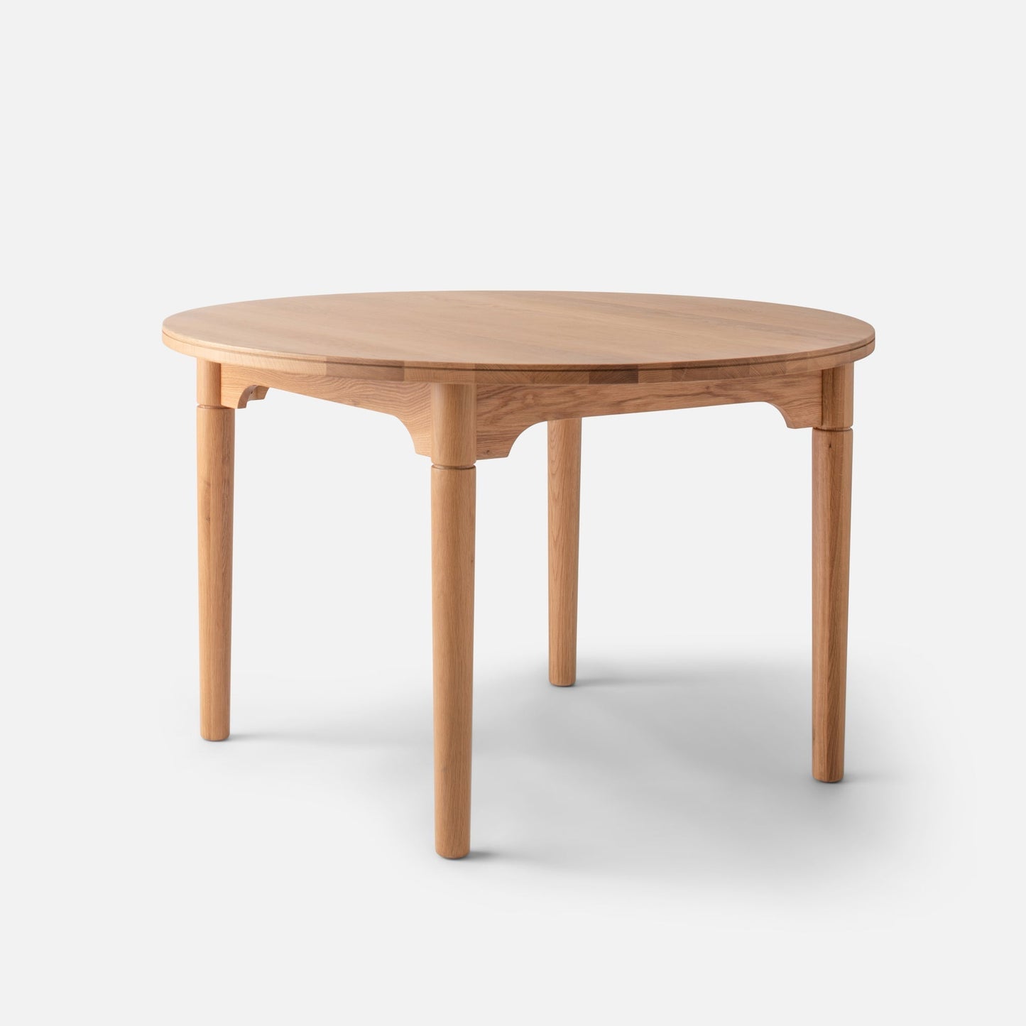 Three-quarter view of Willard Dining Table showcasing round solid oak top, tapered legs, and apron on white background