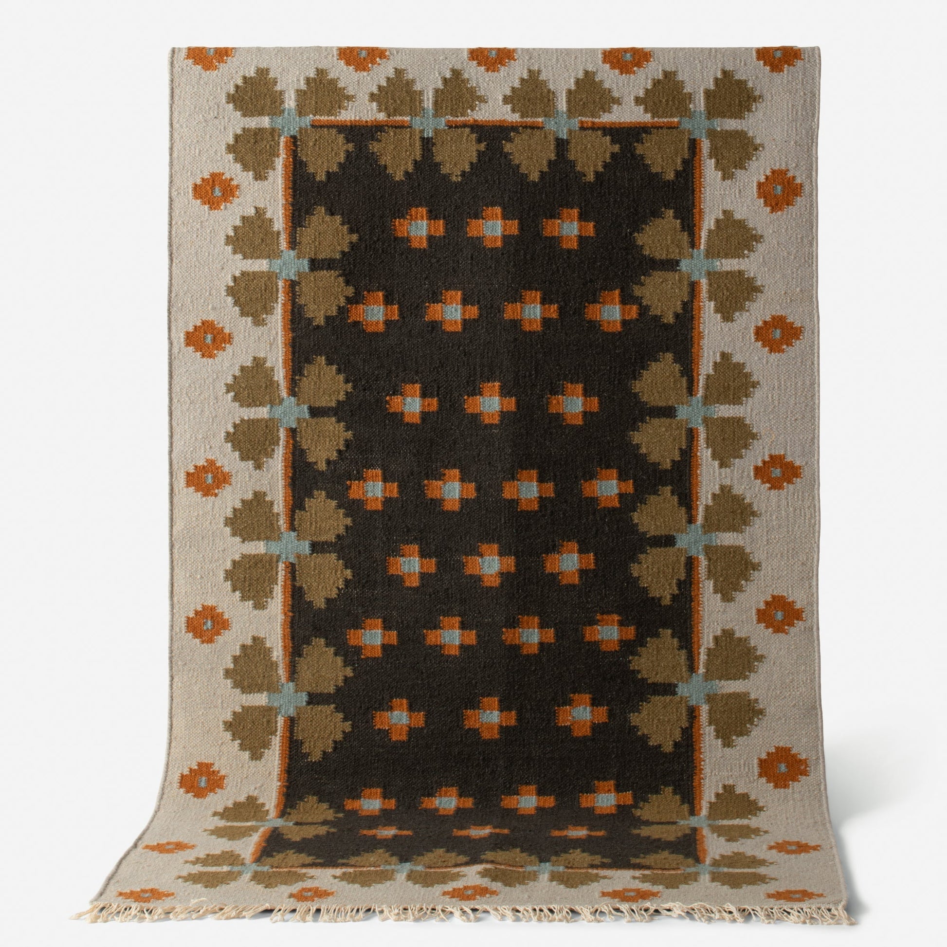 Angled view of Flora Wool Flatweave Rug showcasing geometric Scandinavian folk-floral pattern in black, tan, orange, and blue with fringed edge on white background
