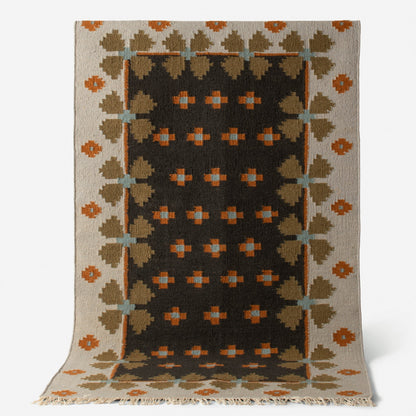 Angled view of Flora Wool Flatweave Rug showcasing geometric Scandinavian folk-floral pattern in black, tan, orange, and blue with fringed edge on white background