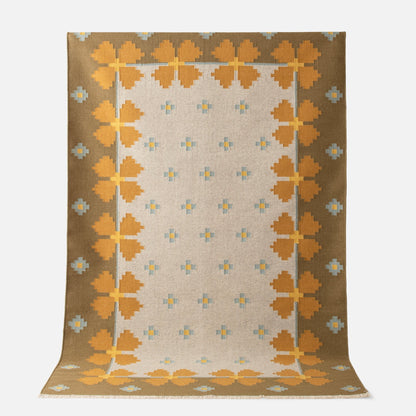 Angled top-down view of Flora Wool Flatweave Rug showcasing golden yellow floral border and small blue cross motifs on a beige field with white background