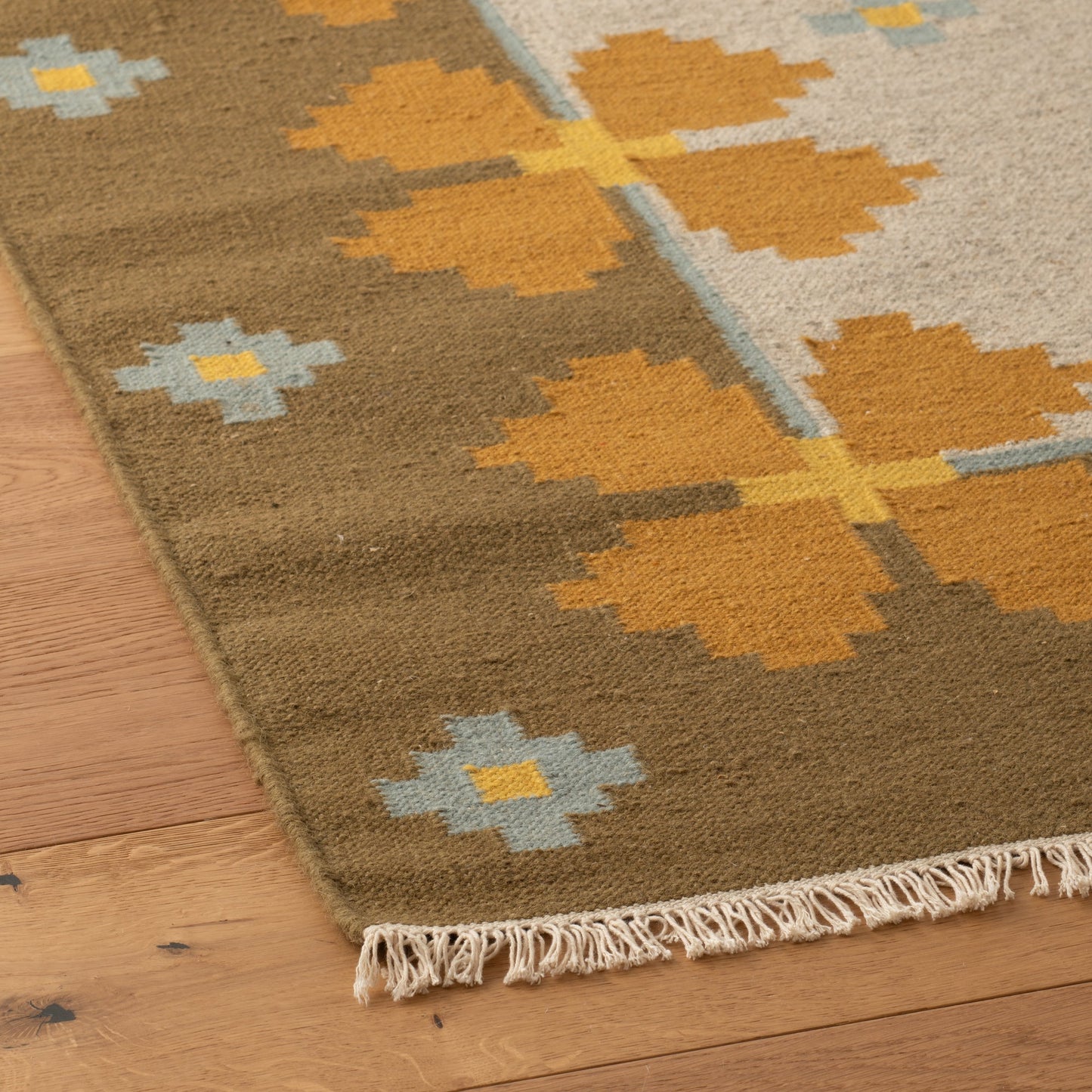 Angled close-up of Flora Wool Flatweave Rug showcasing mustard floral motifs, light blue accents, fringe on a hardwood floor